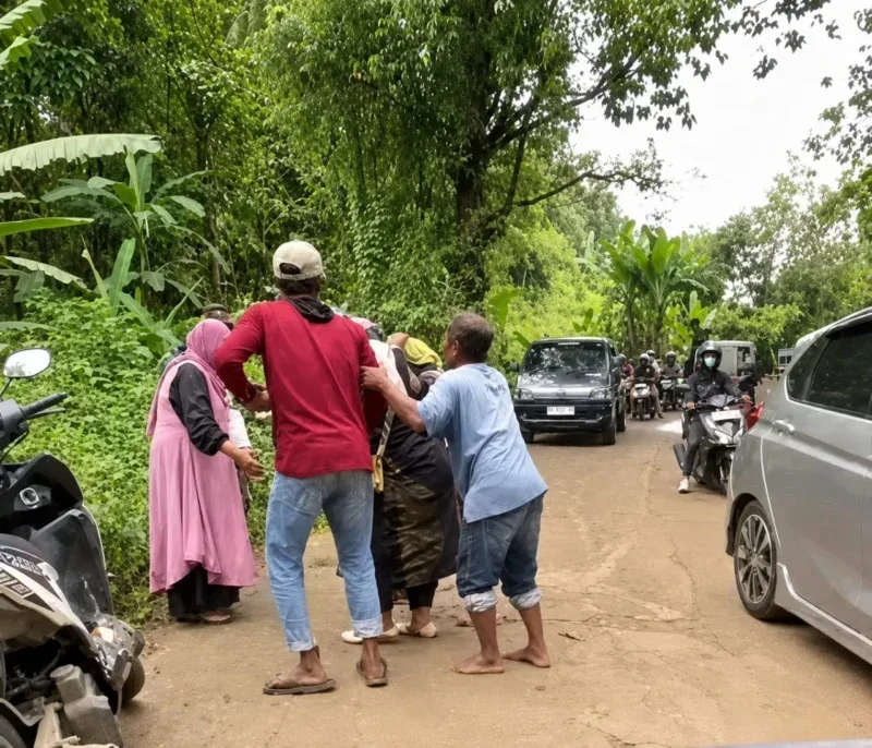 Seorang pemotor terjatuh saat melintasi jalan berlumpur dan licin di kawasan pedesaan, sementara warga sekitar berupaya memberikan pertolongan.